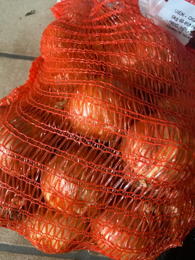 The image shows a close-up view of a mesh bag filled with onions. The bag is bright orange and the onions inside are a pale yellow or white. The mesh is tightly woven, creating a diamond pattern through which the onions are visible. A white label is partially visible at the top right of the bag, with some text on it that appears to be in Dutch or a similar language, including "UIEN - OIG", "5kg 60-80", and "ORIG. HOLL...". The onions appear to be of a medium size. The background is out of focus but seems to be a textured surface, possibly concrete or stone. There are no people or animals visible in the image. The scene is likely outdoors, given the lighting and the background. No specific location in Kassel, Germany can be determined from the image.