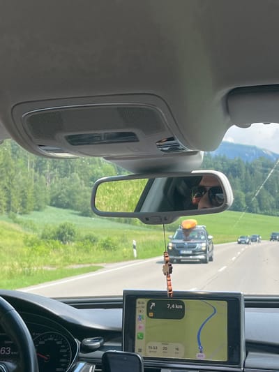 The image shows the interior of a car driving on a road with lush green fields and trees on either side. In the foreground, the dashboard and steering wheel are visible, along with a GPS navigation screen displaying a route. The GPS indicates a distance of 7.4 km remaining and an estimated arrival time of 15:53. A rearview mirror reflects the driver wearing sunglasses. Ahead, several cars are driving on the road, one of which has a kayak or similar item strapped to its roof. The scene appears to be set in Oberammergau, Germany, given the rural, mountainous landscape typical of the Bavarian region. The weather is bright and sunny, suggesting a summer day. The overall mood is one of a leisurely road trip through the countryside.