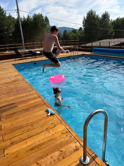 In Elekmonar, Russia, a young male in black shorts is captured mid-air as he jumps into a bright blue swimming pool. He is positioned above and to the left of a woman who is swimming in the pool. To the woman's right, a pink inflatable ring floats on the surface of the water. Nearby, on the wooden deck of the pool, a white and blue water gun rests on the planks. The scene is set outdoors, with a lush forest of tall pine trees bordering the pool area. The sky above is partly cloudy, with a hint of mountains visible in the distance. A wooden fence and structures are also present around the pool.