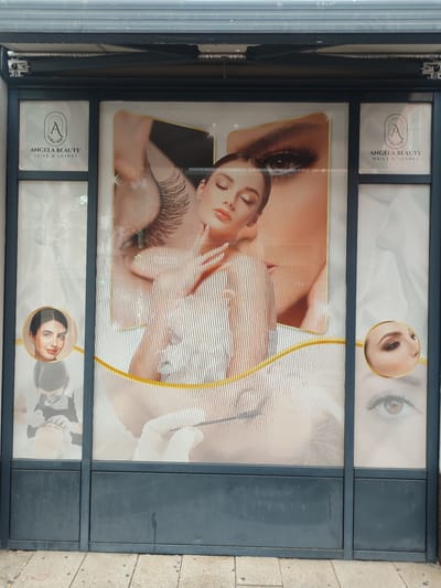 This is a full, eye-level, exterior shot of a commercial storefront in Rosenheim, Germany. The storefront features a large window display with a stylized advertisement for "ANGELA BEAUTY NAILS & LASHES." The advertisement is dominated by close-up images of a woman's face, focusing on her eyes and delicate makeup, as well as hands with manicured nails. The company logo, an 'A' enclosed in an oval, is visible on both the left and right sides of the display. The storefront is framed by dark gray metal panels on the bottom and a dark gray awning above. The display is illuminated from behind, giving the images a soft glow. The sidewalk in front of the store is made of concrete pavers.