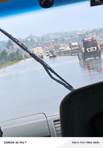 The image presents a wet, multi-lane road scene in Muguga ward, Kenya, captured from within a vehicle looking through its rain-speckled windshield. A dark-colored windshield wiper blade is prominently positioned in the mid-left. The asphalt road is thoroughly wet and reflective, indicating recent or ongoing rainfall, with multiple horizontal white lines marked across its surface.

Traffic is observed moving away from the viewer. In the right midground, a dark-colored SUV, equipped with a roof rack, displays illuminated red taillights that reflect on the wet pavement. A Kenyan license plate, partially visible, shows characters "KSN 097Z." Behind this SUV, a white and yellow passenger van (matatu) proceeds in the same direction. Further into the midground and left, several large commercial trucks (lorries) are visible.

The background features undulating hills densely covered with buildings of varying sizes and interspersed with green vegetation under an overcast sky. The immediate foreground includes a portion of a dark headrest or seat in the lower right and a light-colored vehicle dashboard section with vents in the lower left. A small dark device is mounted on the top center of the windshield, near the blue tinted strip. No pedestrians or explicit human figures are discernible, though drivers are implied within the vehicles.