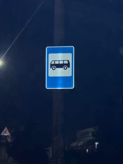 A dark scene featuring a blue and white rectangular road sign with a bus icon displayed vertically. The sign indicates a bus stop or bus lane. In the lower right corner, a faded triangular sign is visible. The background is dark, suggesting nighttime, with a building partially visible on the right side. A bright light source reflects off the road surface and the sign.