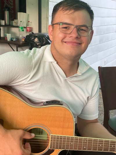 A young man with short dark hair and glasses is playing an acoustic guitar. He is wearing a white polo shirt. The guitar has a light-colored wooden body with a soundhole that reveals a green label with "YAMAHA FGX820C" printed on it. His right hand is strumming the strings. In the background, a brown chair is visible. The scene appears to be outdoors, with a white-paneled wall and some greenery in the distance. There are also some items on a shelf in the background, including bottles of soda and cups. The image was taken in Beltershain, Germany.