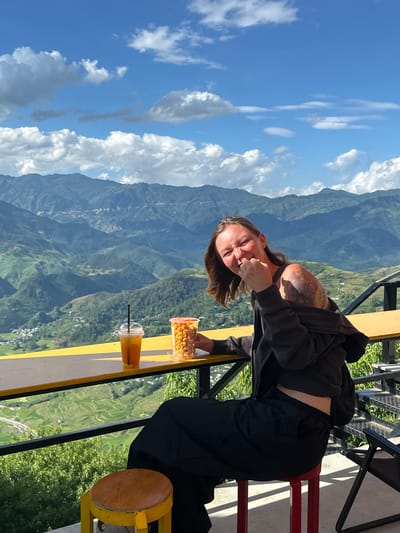 An adult female is seated at an outdoor establishment overlooking a vast mountainous landscape under a blue sky with scattered white clouds, consistent with the terrain found in Tu Le, Vietnam. She is positioned at a high yellow table or counter, facing slightly left towards the camera, and displaying a broad smile or laugh with her eyes squinted. Her right hand is raised to her mouth, holding a clear plastic cup filled with orange-colored popcorn. A clear plastic cup with a black straw, containing an amber beverage with fruit slices, rests on the table to her left. She wears a dark zip-up hoodie, partially pulled off her left shoulder, revealing extensive tattoos on her left shoulder and upper arm. She also wears a dark top, showing a sliver of her midriff, and dark trousers. She is seated on a red stool; another yellow-legged, wooden-topped stool is partially visible to the far left under the table. A black metal railing runs below the yellow table. The background features multiple layers of green, forested mountains with valleys, where scattered clusters of buildings and visible terraced fields indicate human settlement. The sunlight is bright, casting distinct shadows.