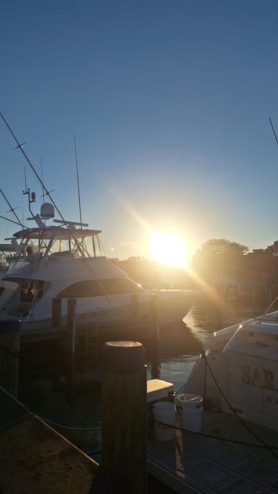 The image depicts a serene harbor scene in Nantucket, Massachusetts, USA, illuminated by the warm glow of a setting sun. In the foreground, sturdy wooden pilings anchor a dock. Moored boats, including a large white yacht with fishing poles prominently displayed, occupy the middle ground. The yacht's name, "SARAH," is partially visible in golden lettering on its stern. The water reflects the sky and the boats, creating a tranquil atmosphere. Trees and a building line the distant shore, silhouetted against the vibrant orange and yellow hues of the sunset. The sky is a clear, deep blue, transitioning to lighter tones near the horizon where the sun is setting. Two white buckets sit on the dock near the stern of the "SARAH." The overall impression is one of peacefulness and the end of a day by the water.