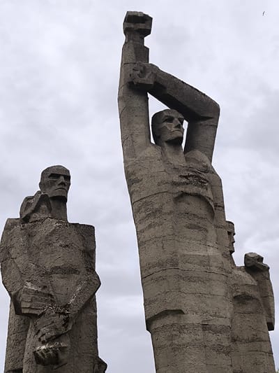 The image depicts a sculptural composition in Mežezeri, Latvia. The sculptures, made of a textured concrete material, represent stylized human figures. On the left are two figures standing close together. The figure on the right raises both arms above its head with hands clenched. The background is filled with a cloudy sky.