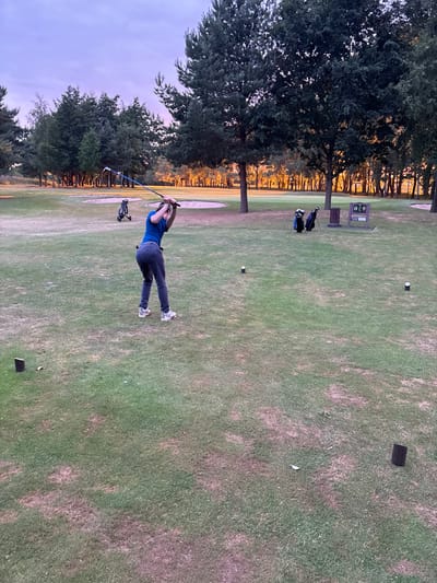 A man in a blue shirt and dark pants is swinging a golf club on a green golf course. His body is angled slightly towards the camera. Behind him, there is a line of trees with some foliage and a glimpse of a sunset. A golf cart is parked in the distance. There are scattered golf tees and golf bags visible on the course, along with a sign board. The grass is mowed and somewhat patchy.