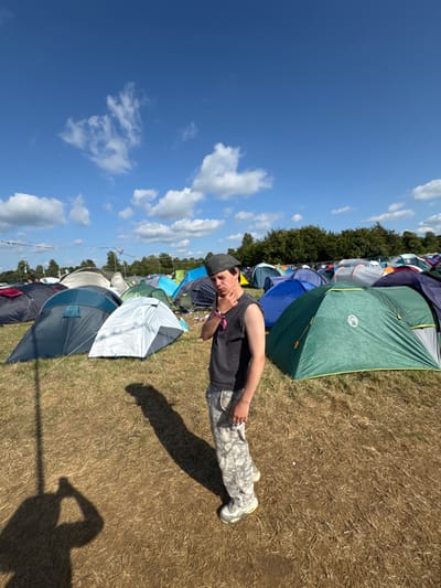 Under a bright blue sky dotted with white clouds, a young person lies on dry, grassy ground amidst a colorful array of tents. The tents range from green and blue to white and grey, creating a vibrant camping scene. The person wears a dark sleeveless shirt, patterned pants, and light-colored shoes, casting a distinct shadow on the sun-baked earth. A wristband adorns their left wrist, suggesting they're attending a festival or event. The backdrop of tents extends into the distance, marked by a line of trees.