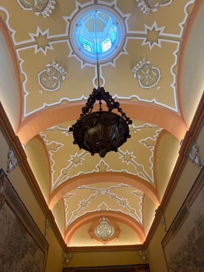 This is a low-angle, interior shot of a hallway ceiling in the Alcázar of Seville, Spain. The ceiling is ornately decorated with a yellow background, accented by white stucco detailing and orange curved beams. A large, dark, antique chandelier hangs from the center, suspended by a chain. In the distance, a circular stained-glass skylight is visible. Along the walls, large tapestries are displayed. The overall impression is one of grandeur and historical richness.