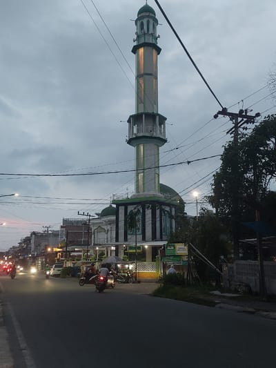 This media file captures a street view in Pematang Siantar, Indonesia, during what appears to be dusk or early evening. The central subject is a mosque with a tall minaret, adorned with green and white accents and illuminated by warm lights, suggesting it is a place of worship. The minaret is a prominent vertical element against a cloudy, overcast sky.
The scene depicts a street with a paved road on the right side of the frame. Several motorcycles are visible on the road, some with their headlights on, indicating ongoing traffic. People are seen riding these motorcycles, suggesting daily commute or evening activities.
The surrounding environment includes buildings, some with illuminated windows and storefronts, lining the street. There are also trees and utility poles with a network of electrical wires crisscrossing the frame, common in urban Asian landscapes. A signpost is visible with text that reads "PANCIGAS NASUTUON" in yellow lettering on a green background, likely indicating a business or location name. Another sign with "Gojek" logo is also visible, suggesting ride-sharing services are available.
The overall mood is that of a typical evening in a busy urban area, with the mosque standing as a significant architectural and cultural landmark amidst the daily life. The time of day, indicated by the low light and artificial illumination, contributes to a slightly subdued yet active atmosphere. The weather appears to be overcast, with no direct sunlight.