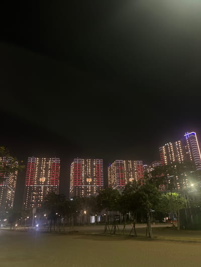 The image captures a nighttime scene in Muara, Indonesia, featuring a row of illuminated skyscrapers. The buildings are lit with a combination of white and red lights, creating a vibrant display against the dark sky. Trees with lush green foliage line the sidewalk in front of the buildings, with some appearing to be newly planted and supported by stakes. A paved walkway or road is visible to the right of the trees, with a faint sheen suggesting it might be wet, possibly due to rain or recent watering. Streetlights are also visible, casting a warm glow on the surroundings. The overall mood is one of urban vibrancy and modern architecture. There is no visible text in the image. The time of day is clearly night, and the weather appears calm. The setting is an urban environment, likely a residential or commercial district in Muara, Indonesia, characterized by contemporary high-rise buildings and landscaping.