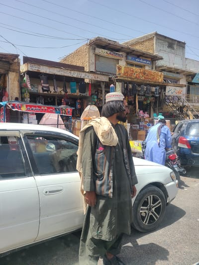 A man stands to the left of a white sedan on a street in Saranan, Pakistan. The man wears a dark green tunic, green pants, a light brown scarf draped over his shoulders, and a white and red patterned skullcap. He has dark hair and a beard. Behind the man, shops line the street, displaying clothing, toys, and other goods. A pink tent is visible in one of the shops. A person in a light blue tunic walks by a motorcycle, and a dark blue car is parked to the right. The sky is clear and blue. The street is paved with asphalt.