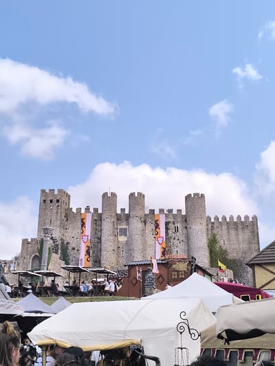 The image depicts a vibrant scene outside the Óbidos Castle in Óbidos, Portugal, under a partially cloudy blue sky. The castle, an imposing medieval stone structure with multiple turrets and battlements, dominates the background. Two large banners hang from the castle walls, one featuring a dragon and a coat of arms, and another with a dragon graphic. In front of the castle, a bustling outdoor market is in full swing, with numerous stalls and tents set up. People are seated at picnic tables under shaded awnings, enjoying a meal or drinks. Another section of the market features tents and stalls, some with draped fabric and decorative elements. A statue of a gargoyle is visible on a stone structure. In the foreground, more tents and a crowd of people suggest a lively event or festival. The overall impression is one of a historical reenactment or cultural fair taking place at a significant Portuguese landmark.