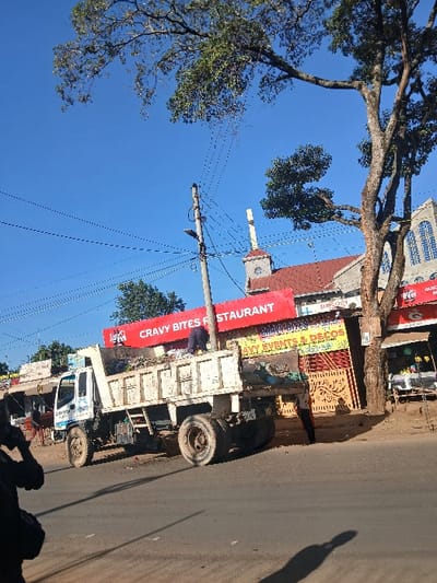 The image captures an outdoor street scene in Hospital ward, Kenya, under a clear blue sky with bright sunlight. A white dump truck, bearing visible text on its cab, is parked on the paved roadside, its bed containing a load of assorted refuse or debris. One individual, dressed in dark clothing, stands within the truck bed, positioned towards the front. A second individual, wearing lighter attire, stands on the dirt shoulder immediately behind the truck. In the far left foreground, the partial silhouette of a third person, appearing to hold a phone to their ear, is visible.
A prominent wooden utility pole with multiple overhead power lines and attached street lights stands to the right of the truck. In the background, a series of buildings line the street. A red sign with white lettering clearly displays "CRAVY BITES RESTAURANT" above "CRAVY EVENTS & DECOS." To the far right, partially obscured by a large tree with green foliage, a building with a red tiled roof, arched windows, and a steeple-like structure topped with a white cross is visible. Shadows from trees and unseen objects are cast across the paved road surface.