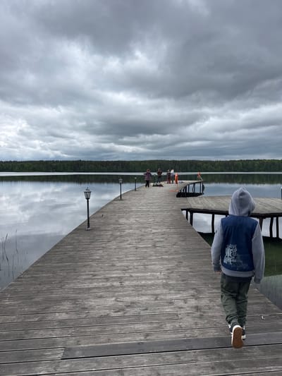 A child in a hooded sweatshirt, dark pants, and sneakers stands on a wooden dock, looking out towards a lake. Other people stand further down the dock, near boats. The sky is cloudy. The location is likely Aleksandrovka, Russia.