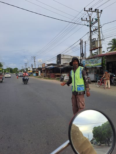 A street scene unfolds in Gampongbaro, Indonesia, under an overcast sky. A man, wearing a black hat and a high-visibility lime green vest with blue checkered accents, stands in the middle of the road, his mouth slightly open as if speaking or whistling. He is also holding a cigarette in his mouth and has one hand extended, palm up, as if signaling. His attire includes a patterned long-sleeved shirt and brown cargo pants.

The road appears to be a two-lane asphalt surface with traffic. Several motorcycles are visible, some with riders. On the right side of the road, a red bicycle leans against a man wearing a patterned shirt and a blue cap. Behind them, various small shops and buildings line the street, some with awnings and signs. Electrical poles with a tangle of wires are prominent overhead. In the foreground, the circular reflection in a motorcycle's rearview mirror shows a glimpse of trees and the sky, with a hand visible in the lower portion of the mirror. The overall atmosphere suggests a typical day on a local road.