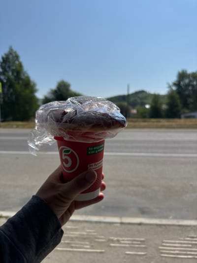 The image shows a person holding a red paper cup with a baked good wrapped in clear plastic. The cup has a logo with the number "5" and some text in Russian. The text on the cup translates to "For taste, like in childhood" and "For you to enjoy." The person's hand is visible, wearing a grey sweatshirt sleeve. In the background, there is a paved area with white lines, possibly a road or sports track, and some trees and a clear blue sky. The scene appears to be outdoors on a sunny day. The orientation of the image is landscape. The general setting suggests an outdoor commercial area, perhaps near a food vendor or a store in Mayma, Russia. The overall mood seems casual and everyday.