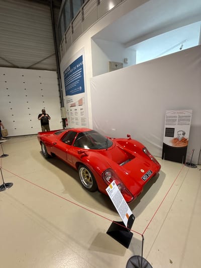 A red McLaren F1 LM sits within a museum exhibit, cordoned off by a red rope barrier. The car's license plate reads "NOV 2105". Informational plaques are displayed near the car, including one with a portrait and text in English. A man is visible in the background near a white wall with a large sign that reads "Le Musée des 24 Heures du Mans". The ceiling is high and white, with natural light coming in.