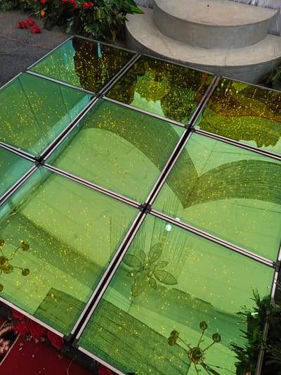 In Meuliek, Indonesia, a close-up aerial shot captures a section of a green glass dance floor illuminated with small yellow lights, possibly LED, embedded within or beneath the surface. The dance floor is divided into a grid of nine square panels, each outlined with metallic silver framing. Reflections on the glass reveal a blurred overhead scene, suggesting an event or gathering, with hints of floral arrangements and architectural elements like a tiered, semi-circular stage. The floor itself displays a dappled pattern of lights and abstract shapes, possibly from the underlying illumination or decorative elements. In the lower left corner, a portion of a red carpet with a dark pattern is visible, along with scattered red rose petals, indicating a festive or ceremonial setting. The overall impression is one of a decorated event space.