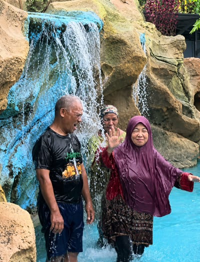 In Eampat Kwalamencirim, Indonesia, three individuals are standing in the turquoise water of a pool, partially obscured by a cascading waterfall. The waterfall flows over a textured rock formation painted with streaks of blue and yellow. To the left, a man with graying hair wears a wet black t-shirt with a graphic design and navy blue shorts, his mouth open as if shouting or laughing. Behind him, a person wearing a headscarf and a red garment can be seen. On the right, a woman wearing a purple hijab and a wet red garment over a patterned skirt and black leggings smiles and waves with her right hand. The water is splashing around them, indicating they are actively interacting with the waterfall.