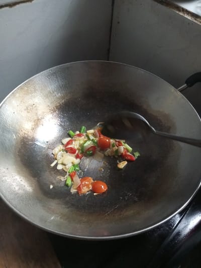 In a kitchen in Ariyalai East, Sri Lanka, a dark, well-used metallic wok contains a mixture of sautéing ingredients. The ingredients include chopped white onions or shallots, green scallions, small red cherry tomatoes, and sliced red chilies, all glistening with oil at the bottom of the wok. A dark metallic slotted spoon is positioned over the ingredients, indicating active stirring. The wok rests on a dark stovetop or countertop, with a dark wooden surface visible to the lower left. The background features a light-colored kitchen wall, showing minor discoloration and a corner joint. No people are visible in the scene.