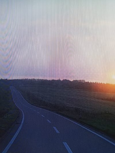 This media file is a photograph taken from a high vantage point, possibly a drone or airplane, overlooking a landscape. The scene is divided diagonally, with the left half dominated by a bright, hazy sky transitioning into soft pink and purple hues near the horizon. The right half features a dark, wooded area with a winding road cutting through it. The road is paved and marked with white lines, indicating it's a travel route. The sun is setting or rising, casting a warm orange glow on the upper right portion of the sky and highlighting the edge of the trees. The overall mood is tranquil and scenic, suggesting a peaceful rural setting at dusk or dawn. There is no visible text in the image. The location is identified as Chachersk, Belarus.