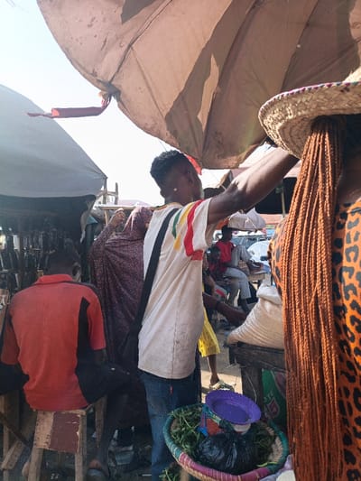 The image depicts an active open-air market scene in Kawaji, Nigeria, under bright sunlight, featuring multiple individuals and temporary fabric shelters. In the immediate foreground right, a person with long, reddish-brown braided hair wears a straw hat and an orange leopard-print garment. Next to them, a male subject, seen from the back, wears a light-colored t-shirt with vertical red, yellow, and green stripes, blue jeans, and a black crossbody bag; his right arm is extended upward towards a large, worn beige fabric umbrella providing shade. A woven basket at his feet contains green leafy vegetables, other small green items, a black plastic bag, and a purple plastic bowl. To the left of the central male, a person in a dark purple patterned garment is partially visible, also with an arm raised. Further left, a person sits on a wooden stool, viewed from the back, wearing an orange t-shirt with black panels and dark shorts. In the background, several other individuals are visible among additional market stalls and coverings, including one person in a red shirt and a large white sack on a wooden table. The ground is unpaved, and a vehicle is partially discernible in the far distance.