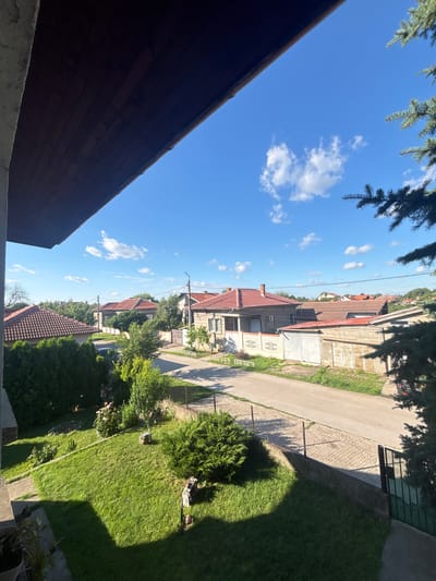 The image depicts a residential street in Marten, Bulgaria, on a clear day. The view is from a slightly elevated position, possibly a balcony or window, looking down at a paved road lined with houses and gardens. The architecture suggests a suburban setting, with many houses featuring red-tiled roofs. Green trees and manicured lawns are prominent in the foreground and along the street. The sky is a vibrant blue with scattered white clouds. There are no people visible in the scene. The overall impression is one of a peaceful, sunny day in a quiet neighborhood.