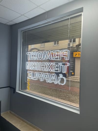 The image shows a neon sign in the window of a shop in Rivne, Ukraine. The sign is illuminated with white neon lights and appears to be in a vertical orientation, with three lines of text and an apple logo at the bottom. The text is in a foreign language, likely Ukrainian, and reads "РЕМОНТ ТЕХНІКИ APPLE" which translates to "REPAIR OF APPLE TECHNOLOGY". The shop is on a street, and the window reflects the opposite building, which has a yellow sign with a bee logo and "B" on it. It is raining, as indicated by the scattered water droplets on the pavement and the reflections in the window. The interior of the shop is not clearly visible due to the reflection and the bright neon sign. The exterior wall of the shop is dark gray, and there is a tiled floor in the foreground.