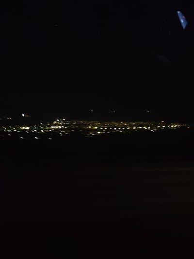 The image shows a nighttime view of a city skyline, likely taken from an elevated position. The scene is dominated by the darkness of the night.  A line of blurred, yellow and white lights defines the city's outline against the black sky. The foreground is black, implying the vantage point is looking out over an area without lights. Given the location of Moshtanets, Bulgaria, the image could potentially be a view of a nearby urban area from a hilltop or high-rise. The photo's quality is low. There are no people visible.