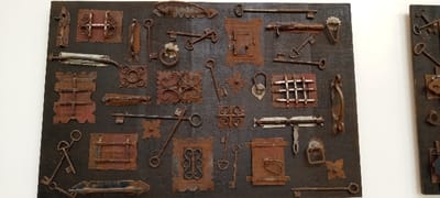 The image depicts a large, dark rectangular panel showcasing an array of aged metallic objects. These objects, primarily composed of rusted iron and other metals, are systematically mounted in a grid-like arrangement across the panel. Identifiable items include numerous keys of varying designs and sizes, diverse lock mechanisms, several door handles (both fixed pull-bar and ring types), assorted latches, bolts, and decorative metal escutcheons and grilles, many exhibiting intricate scrollwork or geometric patterns. The prevalent coloration of the metal components ranges from reddish-brown to dark brown due to oxidation, with some lighter metallic accents. The display panel is positioned against a plain white wall in an indoor setting. A segment of a second, analogous display panel is visible on the right side of the frame. This scene is situated in the city of Yola, Nigeria.
