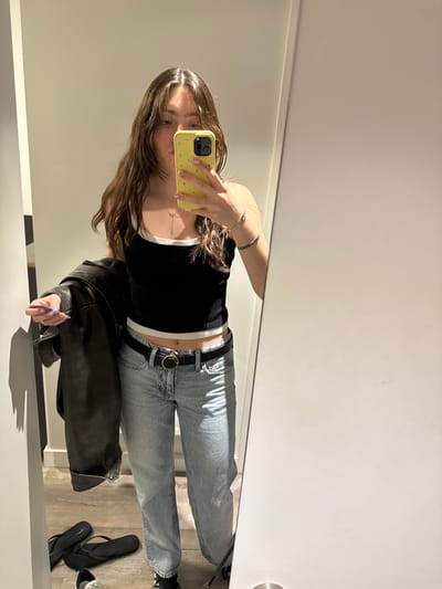 A young woman takes a mirror selfie. She is wearing a black tank top with a white trim, light blue jeans with a black belt, and appears to be holding a black leather jacket. She is holding a yellow smartphone in her hand. Her hair is long and light brown. A pair of black flip-flops are visible on the floor. The setting appears to be a fitting room in a retail store. It is not possible to determine if the location is in Hull, United Kingdom from the image alone.