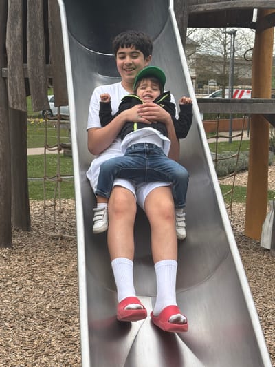 Two people are on a slide. An older boy is lying flat on his back, and a younger child sits on top of him. Both people are smiling. The older boy wears white socks, red slip-on shoes, and white shorts with a white t-shirt. The younger child wears sneakers and a green baseball cap. The slide is made of metal and surrounded by wooden beams. The ground surrounding the slide is covered in mulch. The location of the scene is an outdoor playground, likely in the city of Olingen, Luxembourg.
