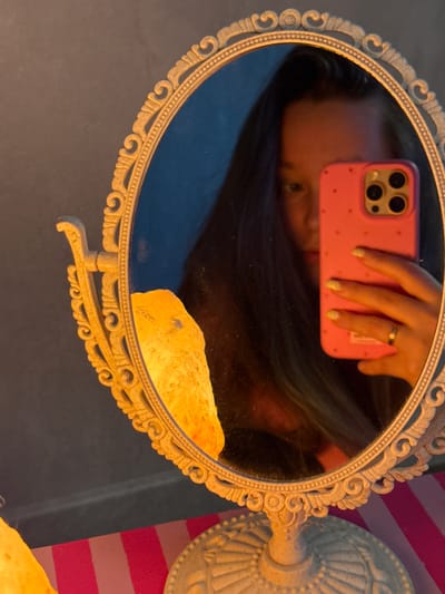 This media file is a close-up, angled shot of a person's reflection in an ornate, oval vanity mirror. The mirror is framed with a cream-colored, decorative border that features swirling scrollwork and a beaded edge. The mirror is tilted, and the reflection shows the upper body of a person, with their face partially obscured. They are holding a pink smartphone with a studded pattern and a prominent camera bump in front of them, as if taking a selfie. A gold ring is visible on one of their fingers, and their fingernails appear to have a glittery polish. The person's dark hair falls around their shoulders.
In the reflection, a light-colored, rough-textured object, possibly a salt lamp or a large crystal, is visible in the lower right portion of the mirror. The background of the reflection is dark and indistinct, but behind the mirror and to the right, a brightly striped pattern of pink and white can be seen, suggesting the presence of decorative wallpaper or fabric. The overall lighting is warm and somewhat dim, possibly indicating an indoor setting in the evening or a dimly lit room. The scene conveys a sense of personal reflection and perhaps preparation or a moment of self-capture. There is no visible text in the image.