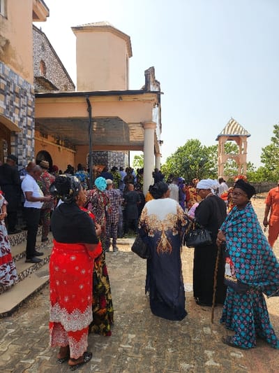 This bright daytime image captures a vibrant outdoor gathering, likely a communal or ceremonial event, in Umuoye, Nigeria. Numerous people, predominantly women, are present, dressed in diverse and colorful traditional African attire. They are standing on a paved ground surface, engaged in conversation or observing the event.
In the foreground, a woman wears a striking red dress elaborately embroidered with silver or white patterns, paired with a black head covering and patterned shoes. Slightly behind her, another woman is seen from the back, sporting a dark blue dress adorned with an intricate white and gold tree-like design across the shoulders and upper back. Further into the crowd, a woman with glasses is visible in a turquoise and black patterned dress, leaning on a dark walking stick. Other attendees are similarly dressed in richly patterned fabrics, reflecting a festive or formal occasion. A man in a white shirt and black trousers is also visible on the left side of the frame.
The setting includes several architectural elements under a clear, bright blue sky. On the left, a light-colored building with a somewhat weathered or unfinished roof is prominent. Towards the bottom left, another structure with patterned tiling adds to the visual texture. In the upper right background, a distinctive, light-colored gazebo-like structure with a patterned roof and arched openings stands out, surrounded by green foliage from distant trees. The ground is paved with interlocking bricks or tiles. The scene conveys a sense of community and celebration. No visible text appears in the image.