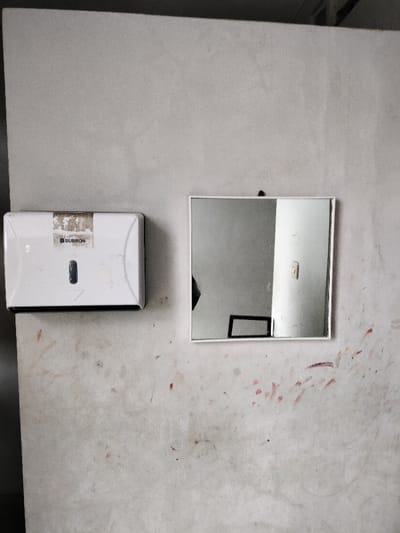 The image displays a light-colored, textured wall with a paper towel dispenser and a square mirror mounted on it. The wall, possibly concrete or painted plaster, shows considerable wear, characterized by numerous smudges, dark marks, and reddish splatters, suggesting a utilitarian or well-used environment.
On the left, a white, rectangular dispenser, likely for paper towels or wipes, is affixed to the wall. It features a visible dark slot. A label on its front is partially peeled and worn, but the brand name "SUBRON" is discernible, accompanied by a small, possibly registered trademark, symbol.
To the right of the dispenser, a square mirror with a simple white frame hangs on the wall. The mirror reflects portions of an interior space. Visible in the reflection are a dark, indistinct silhouette on the left, a rectangular frame (possibly part of a window or furniture) towards the bottom-center, and a bright white vertical structure, perhaps a wall or pillar, on the right side of the reflection. The reflections imply a simple, possibly industrial or functional indoor area.
No specific activity is occurring; the items are stationary fixtures. The overall scene suggests an indoor location, possibly a public restroom, workshop, or utility area, in Dramaga, Indonesia. There are no explicit cues for the time of day, but the lighting is consistent with an indoor setting.