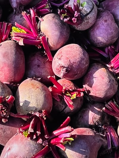 The image is a close-up, overhead shot of a pile of fresh beets. The beets are predominantly a deep reddish-purple color, with some showing lighter, pinkish hues due to lighting. Their skins appear slightly rough and textured. Many of the beets still have their vibrant, fuchsia-colored stems and some small, green leaves attached. The beets are tightly packed together, creating a dense, textured surface. The lighting suggests an outdoor environment, possibly a market or farm stand, with sunlight casting shadows and highlighting the round shapes of the vegetables. There are no people or any indication of the city of Voluntari, Romania, present in the image.