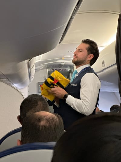 Inside an airplane cabin, a flight attendant in a white shirt and dark vest holds a yellow life vest, demonstrating its use to passengers. The plane's interior, including a grey overhead compartment and an exit sign, are visible in the background. Passengers seated nearby are partially visible in the foreground. The scene suggests a safety briefing or demonstration before takeoff.