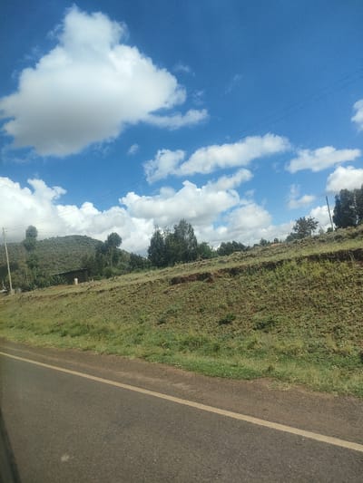 The image shows a rural landscape in Kisima ward, Kenya. A paved road with a yellow dividing line stretches across the foreground. Adjacent to the road, a grassy embankment rises, leading to a plateau with scattered trees and sparse vegetation. A building is visible among the trees. In the background, a hill is covered with trees. The sky is bright blue with scattered puffy white clouds. Overhead power lines are visible.