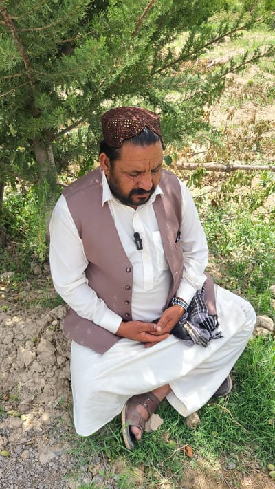 A man is lying down in a grassy area, possibly in the city of Qilla Saifullah, Pakistan. He is wearing a white shalwar kameez, a brown vest, brown sandals, and a brown embroidered cap. A black and white scarf is draped around his neck. The man appears to be resting or sleeping with his hands folded. A small microphone is clipped to his shirt. The environment includes green grass, trees, and a patch of dirt or gravel.