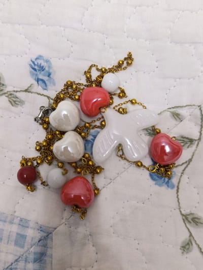 A close-up, overhead shot shows a gold-colored chain with several decorative charms. The charms include white heart-shaped beads, red heart-shaped beads, a white dove-shaped charm, and a single round red bead. The necklace is resting on a white quilted fabric with a subtle floral pattern and some light blue checkered fabric visible in the lower left corner. There is no indication of the city of Semey, Kazakhstan.