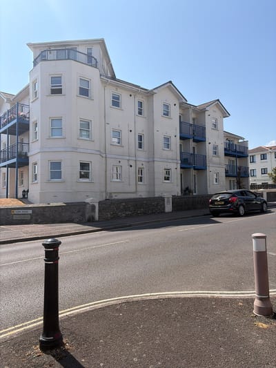 The image shows a street scene in Torquay, United Kingdom, on a sunny day. A modern, pale-colored apartment building with blue balconies dominates the background. A black car is parked on the street in front of the building. A "H YORK ROAD" street sign is visible on a stone wall. Two bollards, one black with red and white stripes and another brown with white accents, are present on the side of the road. The sky is clear and blue.