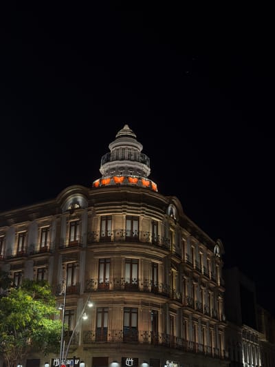 This is a low-angle, night-time shot of an ornate building with a dome and many windows. The building appears to be made of stone with decorative iron balconies. The dome is illuminated with orange light and has a fluted design. The windows are lit from within, casting a warm glow. The facade of the building is richly detailed with columns and arches. In the foreground, lush green trees with illuminated leaves are visible, suggesting a warm climate and possibly springtime or summer. The sky is dark and black, indicating it is nighttime. There are streetlights visible, illuminating the facade and the trees. The overall mood is one of architectural grandeur and nighttime ambiance.

There is visible text on a sign that reads "Ban" and below it "ar". The context of Almería, Spain, suggests this might be a historic or significant building within the city, possibly a hotel, government building, or cultural institution. No people are visible in the image.