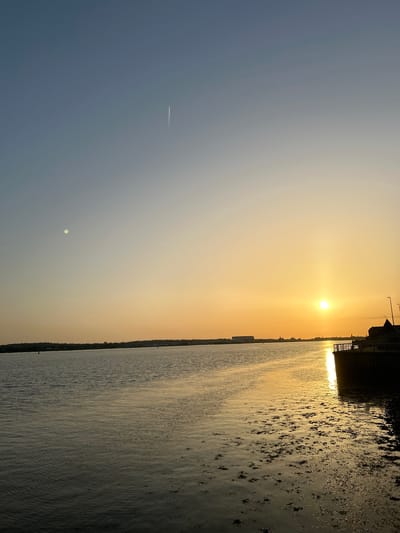 The image depicts a sunset over the water in Liverpool, United Kingdom. The sun is low on the horizon, casting a golden glow across the sky and reflecting on the water's surface. The sky transitions from a pale blue to a warm orange. Silhouetted buildings and structures are visible along the waterfront. The water appears somewhat choppy with patches of seaweed or algae. A contrail can be seen in the sky.
