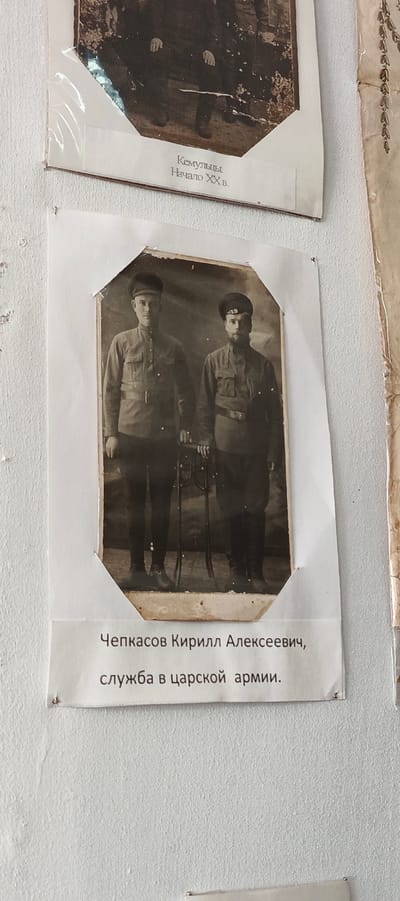 On a textured white wall are three framed black-and-white photographs.
The central photograph features two men in military uniform. The man on the left is clean-shaven with a military cap, a collared shirt, a belted waist, and pants. He is holding a long rifle. The man on the right has a beard and a similar cap. He is wearing the same uniform, with a belted waist. He is holding a rifle. A white label below the photograph says, "Чепкасов Кирилл Алексеевич, служба в царской армии."
The photograph at the top is damaged. It shows a group of people in what appears to be traditional clothing or uniforms from the early 20th century. The label below it says, "Кемульцы. Начало XX в." indicating it is in the city of Кемуль, Russia.
The photograph to the right is partially visible with faint decorative patterns.