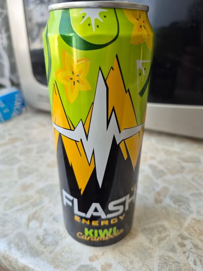 The image shows a close-up of a can of "Flash Energy" drink with a kiwi and carambola flavor. The can is predominantly lime green with yellow and white lightning bolt graphics. The text "FLASH ENERGY" is clearly visible in white, with "Kiwi Carambola" below it in yellow. The can is placed on a speckled, light-colored surface, possibly a table. In the background, out of focus, is a portion of a white appliance, likely a microwave or refrigerator, and a blurred blue object. The scene appears to be indoors. There are no people or animals visible in the image. The lighting suggests it is daytime. The location of Dzerzhinsk, Russia, is not explicitly indicated by any visual cues in the image itself.