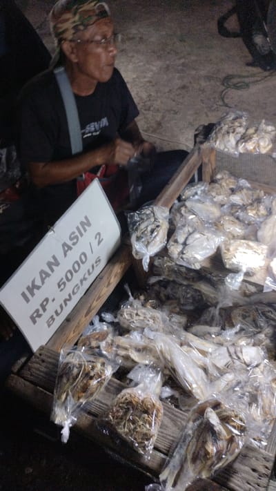 In Sambungrejo, Indonesia, a person wearing glasses and a camouflage bandana sits next to a display of dried fish. The person is wearing a black t-shirt with a gray strap across their chest. They are looking to the right, with a slight smile.
The dried fish are arranged in plastic bags on a wooden crate. Some bags contain whole dried fish, while others contain smaller, shredded pieces of fish. A white sign with text in Indonesian is propped up on the crate. The sign reads "IKAN ASIN RP. 5000 / 2 BUNGKUS," which translates to "Salted Fish Rp. 5000 / 2 Packs."
The scene appears to be outdoors, possibly at a market or roadside stall, given the natural lighting and the informal setup. The time of day is unclear, but the lighting suggests it could be evening or early morning. The overall mood is relaxed and transactional.