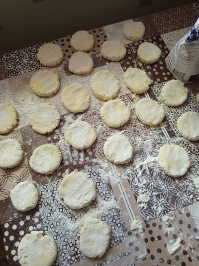 A table covered with a patterned tablecloth is laden with small, circular, uncooked dough patties, dusted with flour. A partially visible bag of flour sits on the right side of the table, with text in Cyrillic. The scene appears to be a kitchen or food preparation area. The image does not provide any information that would allow for a specific location in Novotroitsk, Russia.