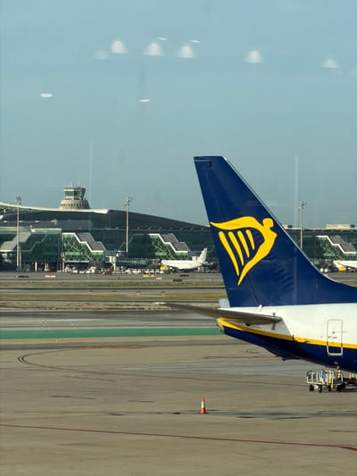 The image shows the tail fin of a Ryanair airplane, prominently displaying the company's distinctive yellow harp logo against a dark blue background. The airplane is on the tarmac of an airport, with the runway and terminal buildings visible in the background. The scene appears to be a sunny day, with clear blue skies reflected on the airplane's fuselage.
The airplane is parked at a gate, with a jet bridge extending towards it, suggesting that passengers may be boarding or disembarking. Other airport vehicles and equipment, such as baggage carts and service vehicles, are also visible on the tarmac. The terminal building is a modern structure with a glass facade, and a control tower is visible on top of one of the buildings.
The location is identified as el Prat de Llobregat, Spain, which is the location of Barcelona-El Prat Airport. The overall impression is one of a busy international airport during daytime operations. The image is taken from a slightly elevated perspective, likely from another airplane or a building overlooking the tarmac. The quality of the image is good, with sharp details and vibrant colors.
