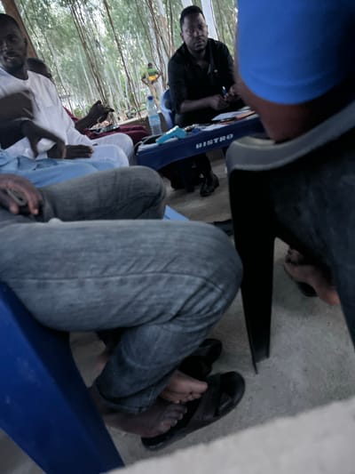 The image depicts a group of people gathered around a blue table with the word "DISTRO" visible on its side. The setting appears to be outdoors, under a canopy of trees with thin trunks, possibly in a park or recreational area. The ground is a light-colored, textured surface, likely concrete or sand.
In the foreground, the legs of two individuals are prominently featured. One person is wearing dark denim jeans and black sandals, with their feet resting on the ground. The other person is also wearing dark denim jeans.
Further back, a man in a white garment is seated, looking to his left. Next to him, another man in a black shirt is seated at the table, holding what appears to be papers or a mobile device. A blue bottle of water and a blue object, possibly a toy or a piece of equipment, are also on the table. In the background, a person in a green and yellow shirt stands near the trees. The overall atmosphere suggests a casual gathering or meeting. The image does not provide specific visual cues to confirm the location as Mayo Dasa, Nigeria, but the presence of individuals who appear to be Nigerian, and the natural environment, are consistent with such a setting.